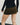 Woman wearing a black minimalist mini skirt that falls mid-thigh, standing confidently. The skirt has a simple, yet elegant design with clean lines and no visible embellishments. She pairs the skirt with a tucked-in white blouse and black pumps for a sophisticated look